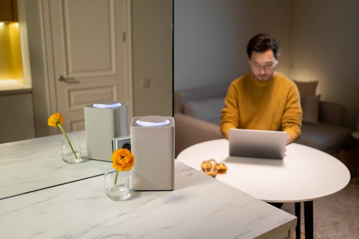 Asian man at a table using a laptop with a smart speaker nearby, indoors.